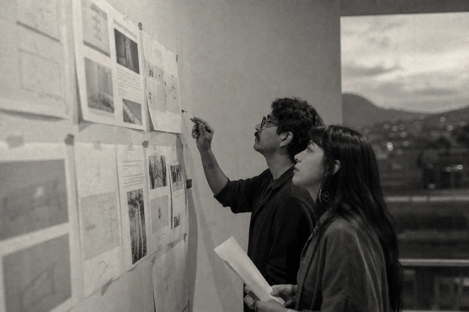 Two people reviewing work pinned to a wall, with a city view, black and white.